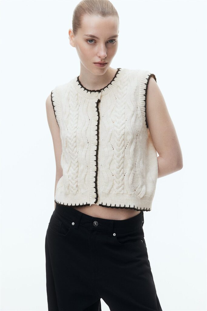 Model wearing a cream cable-knit vest with dark contrast trim.